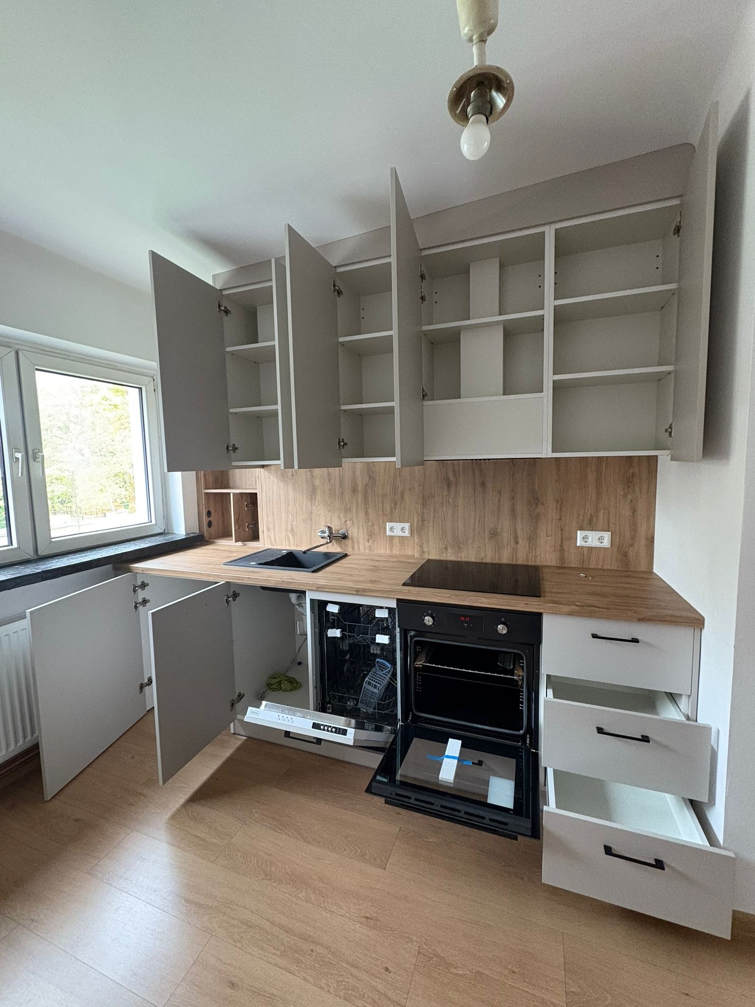 Jasna kuchnia z drewnianym blatem i otwartymi szafkami, ukazująca wnętrze i sprzęt AGD, w tym piekarnik i zmywarkę. Widoczne detale wykończenia i oświetlenie.