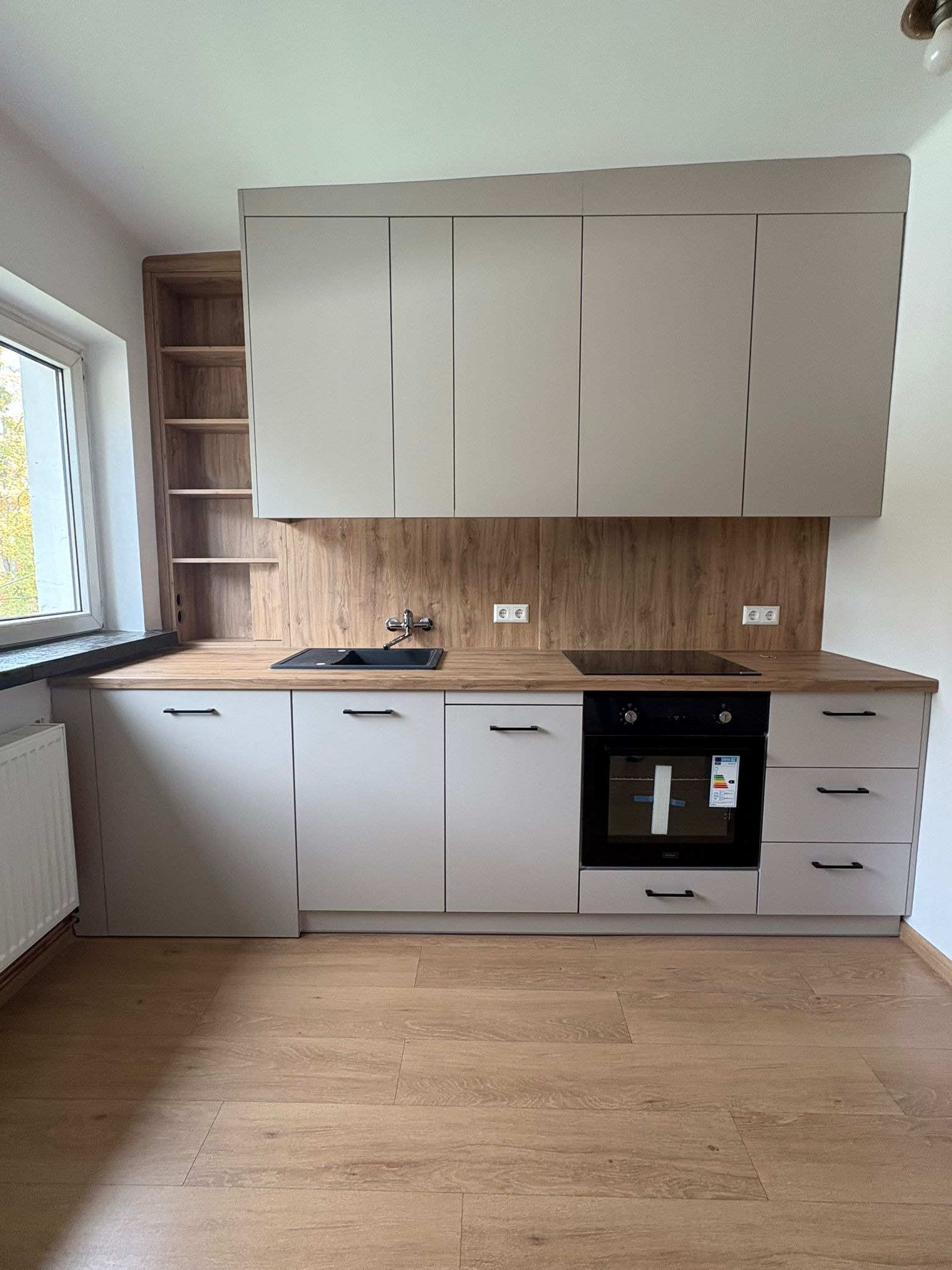 Jasna kuchnia z drewnianym blatem i panelami. Widoczne szafki wiszące i stojące, piekarnik, płyta indukcyjna, zlew i okno. Podłoga wyłożona panelami imitującymi drewno.