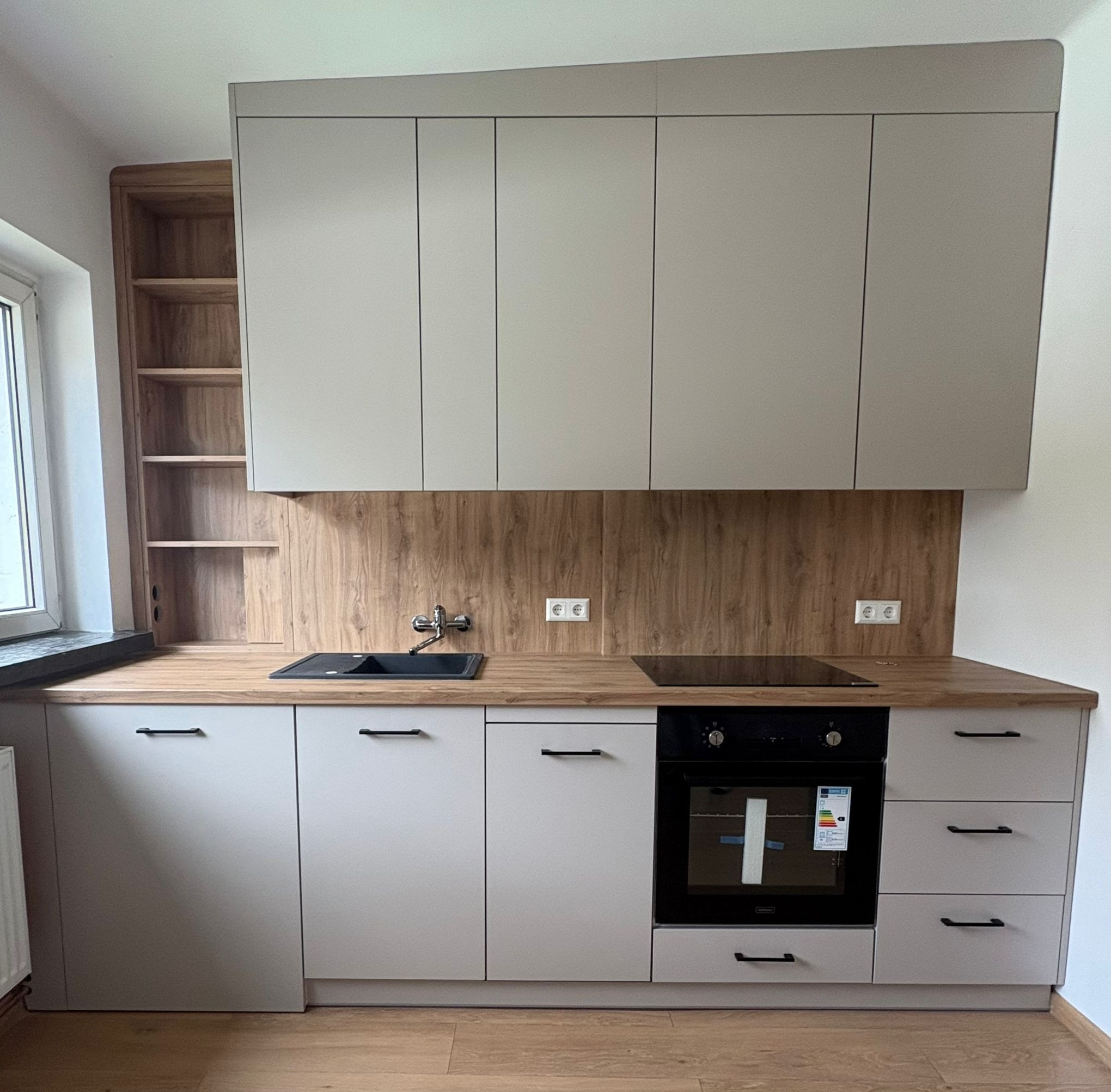 Jasna kuchnia na wymiar z drewnianym blatem i szafkami wiszącymi. Widoczny zlew, płyta indukcyjna i piekarnik. Na ścianie drewnopodobna okładzina. Minimalistyczne uchwyty.