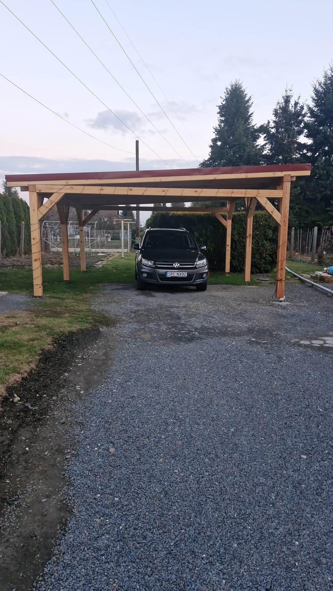 Drewniana wiata garażowa z czerwonym dachem, pod którą stoi czarny samochód. Konstrukcja z belek na żwirowym podłożu, w tle zieleń ogrodu i drzewa iglaste.