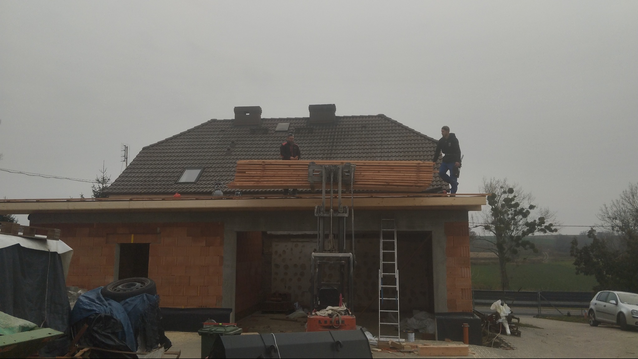 Budowa garażu z drewnianą konstrukcją dachu w trakcie realizacji. Dwóch mężczyzn na dachu układa drewniane elementy. Na pierwszym planie ceglana konstrukcja.