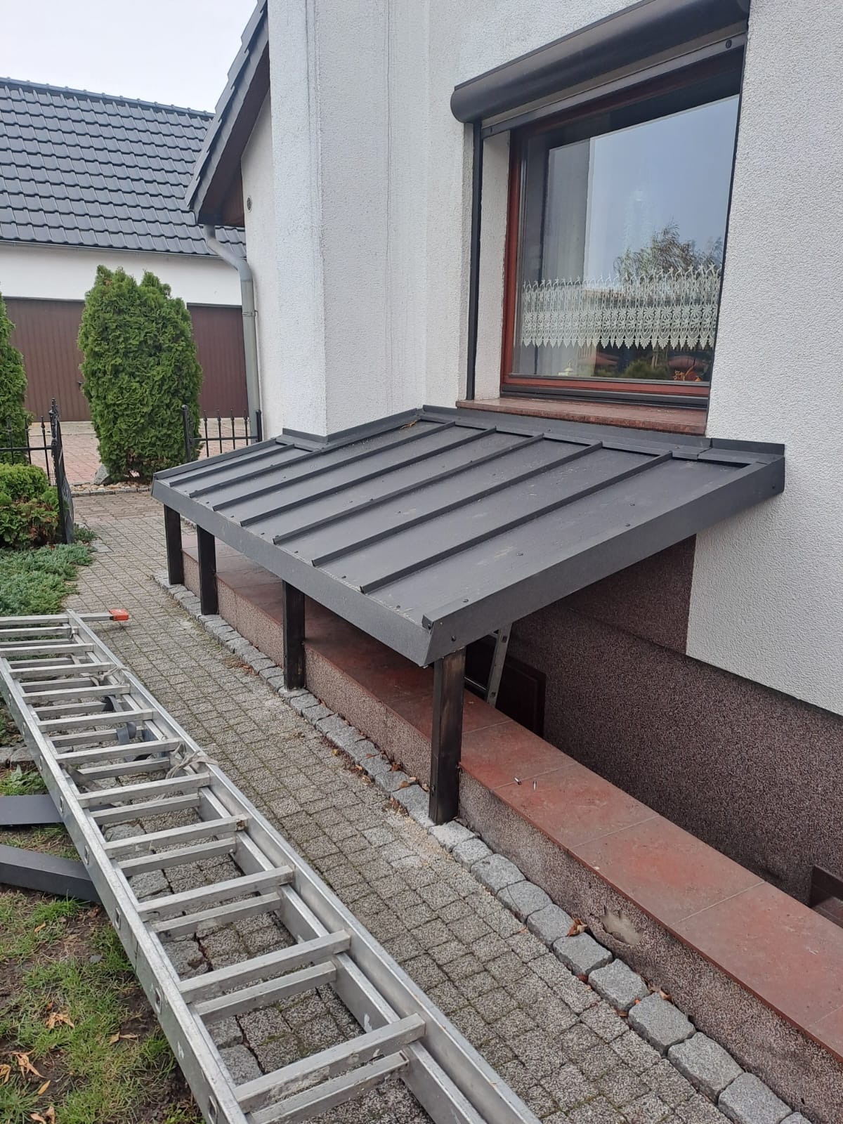 Zadaszenie nad zejściem do piwnicy z szarej blachy trapezowej, wsparte na drewnianych słupkach. Aluminiowa drabina oparta o chodnik z kostki brukowej.