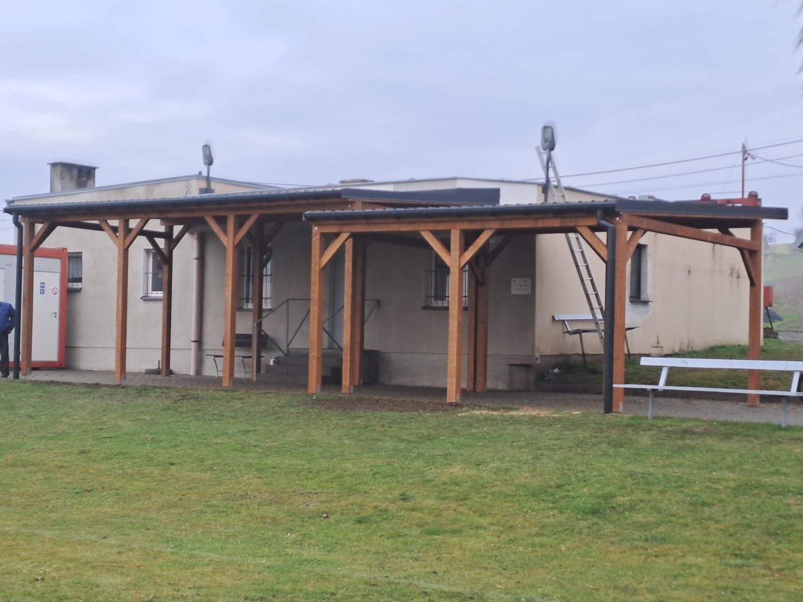 Drewniana pergola z czarnym dachem, przylegająca do budynku z oknami. Na dachu lampa, obok drabina. Z przodu trawnik i biała ławka. Pochmurny dzień.