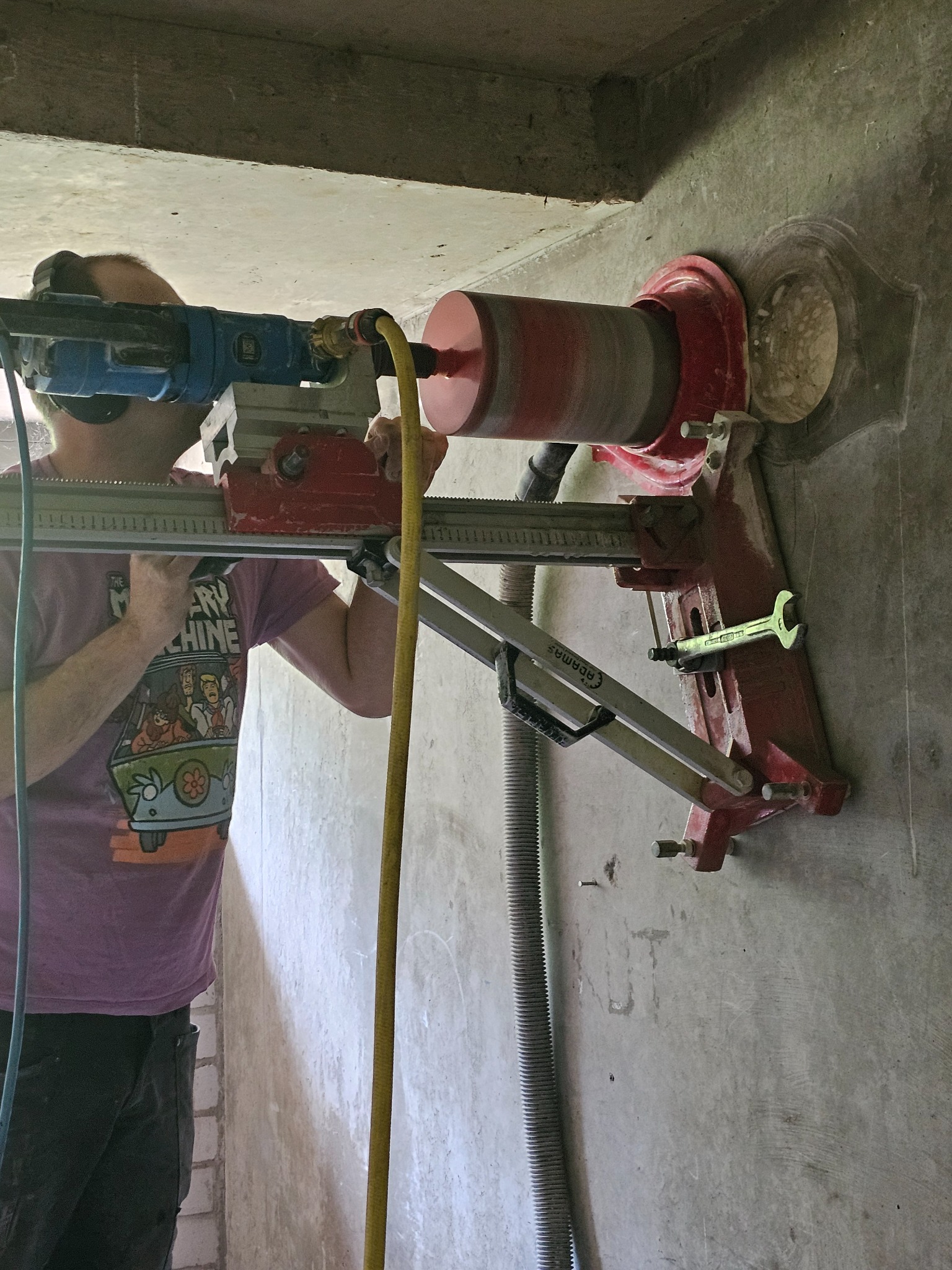 Pracownik w koszulce z motywem Scooby Doo wierci otwór w betonowej ścianie za pomocą wiertnicy rdzeniowej. W tle widoczny fragment instalacji i narzędzia.