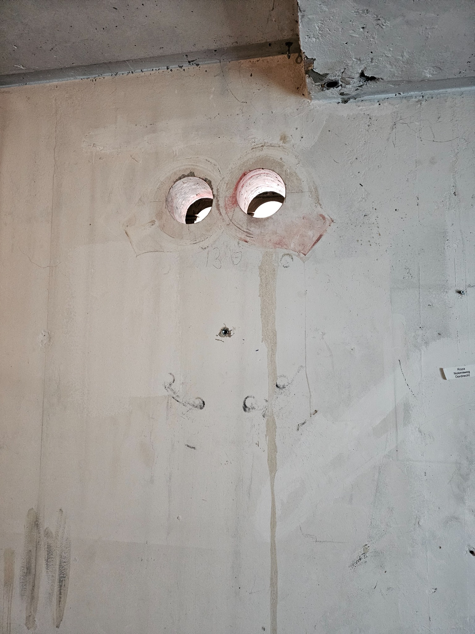 Dwa okrągłe otwory przewiercone w szarej, betonowej ścianie. Widoczne ślady po wierceniu, zarysowania i notatki. Otwory mają czerwone obwódki.