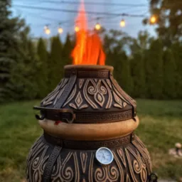 Ozdobny piec ogrodowy typu tandoor z palącym się ogniem wewnątrz, umieszczony na trawniku, z dekoracyjnymi wzorami i termometrem na obudowie.