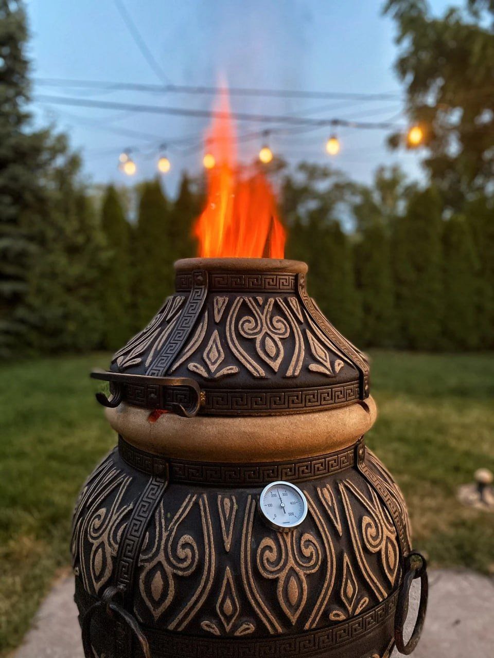 Ozdobny piec ogrodowy typu tandoor z palącym się ogniem wewnątrz, umieszczony na trawniku, z dekoracyjnymi wzorami i termometrem na obudowie.