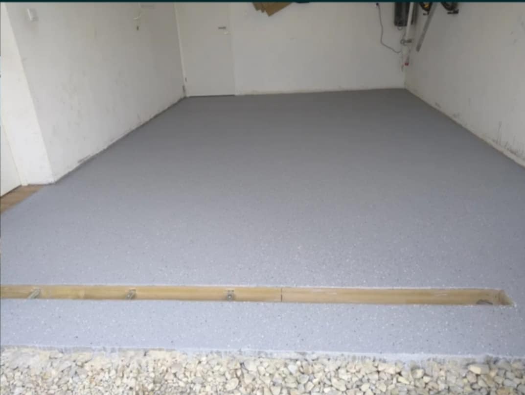 Szara, żywiczna posadzka z odpływem liniowym w garażu w Tychach. Widoczne białe ściany i podłoże z kamyków. Jasne, naturalne oświetlenie.