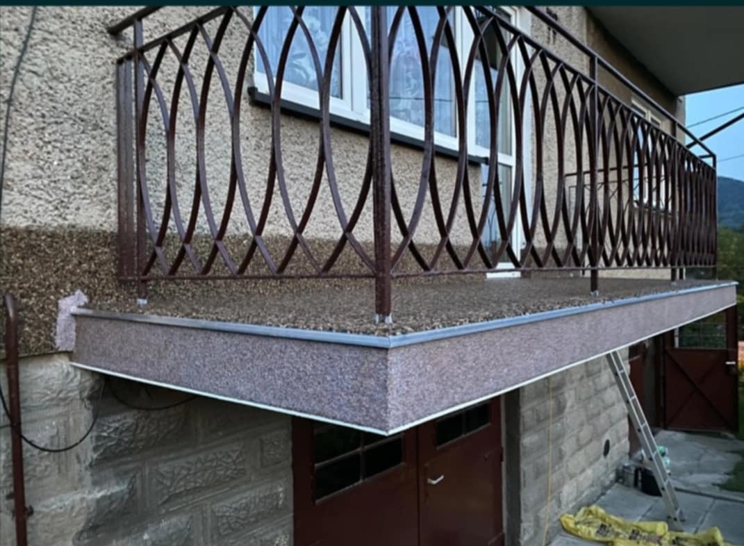 Balkon z ozdobną, brązową balustradą o geometrycznym wzorze, wykończony drobnym kamieniem, z widoczną drabiną opartą o ścianę budynku.