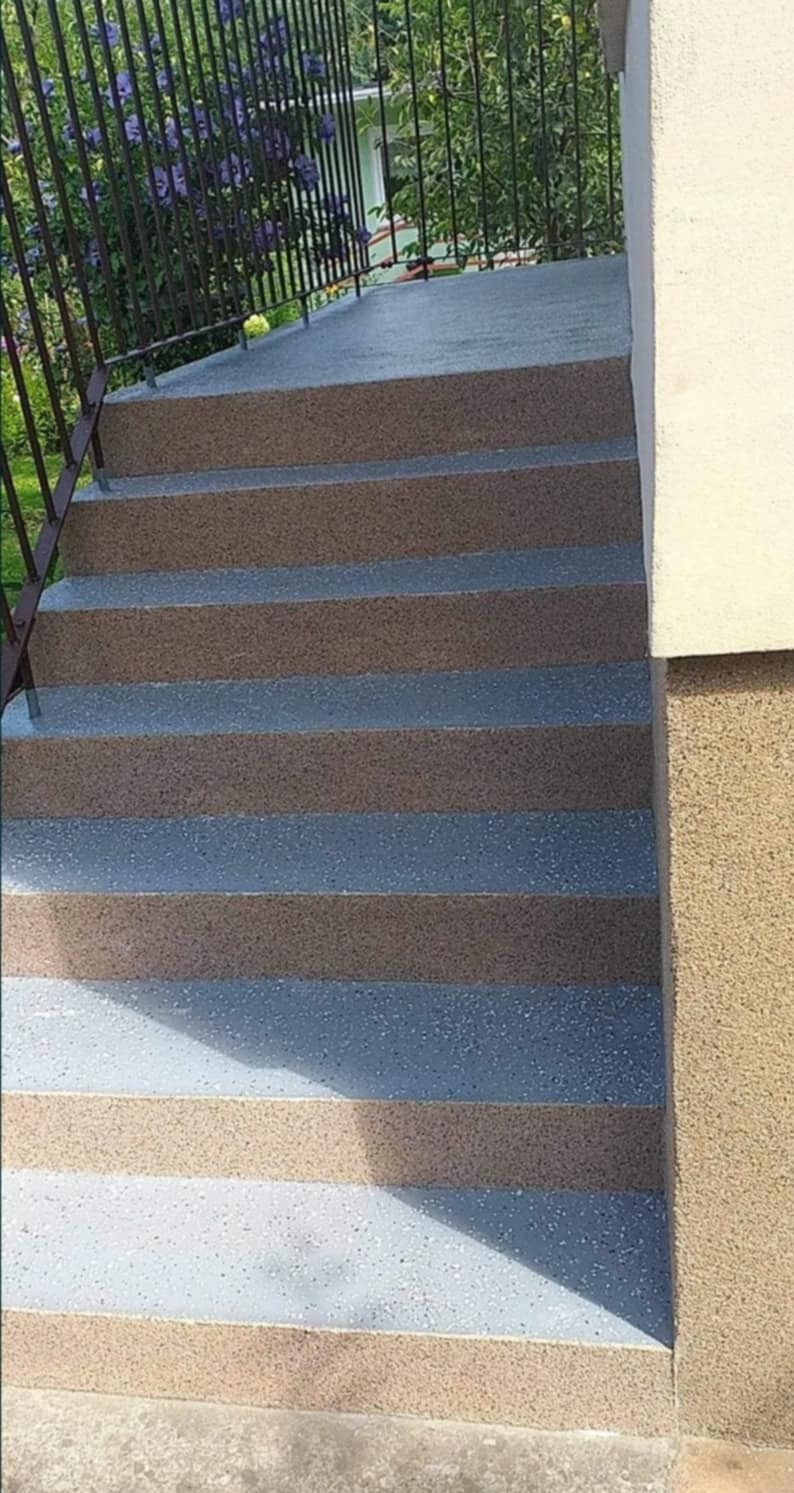 Odnowione schody zewnętrzne z balustradą, pomalowane w naprzemienne pasy beżu i szarości. Widoczne detale faktury i cień.