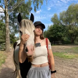 Młoda kobieta w czapce pozuje z koniem na tle drzew i błękitnego nieba. Kobieta trzyma konia za pysk, robiąc zabawną minę. Widoczny telefon w dekolcie.