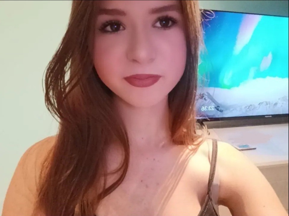 Portret młodej kobiety z brązowymi włosami, z telewizorem w tle, ustawionym na scenę zimową. Ujęcie z bliska, skupione na twarzy.