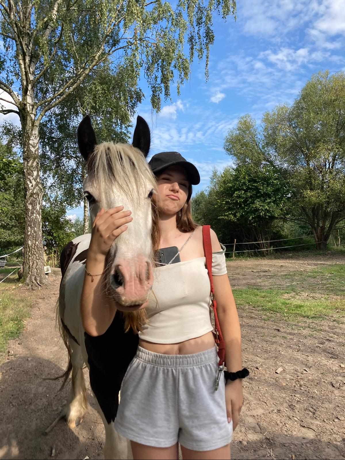 Młoda kobieta w czapce pozuje z koniem na tle drzew i błękitnego nieba. Kobieta trzyma konia za pysk, robiąc zabawną minę. Widoczny telefon w dekolcie.