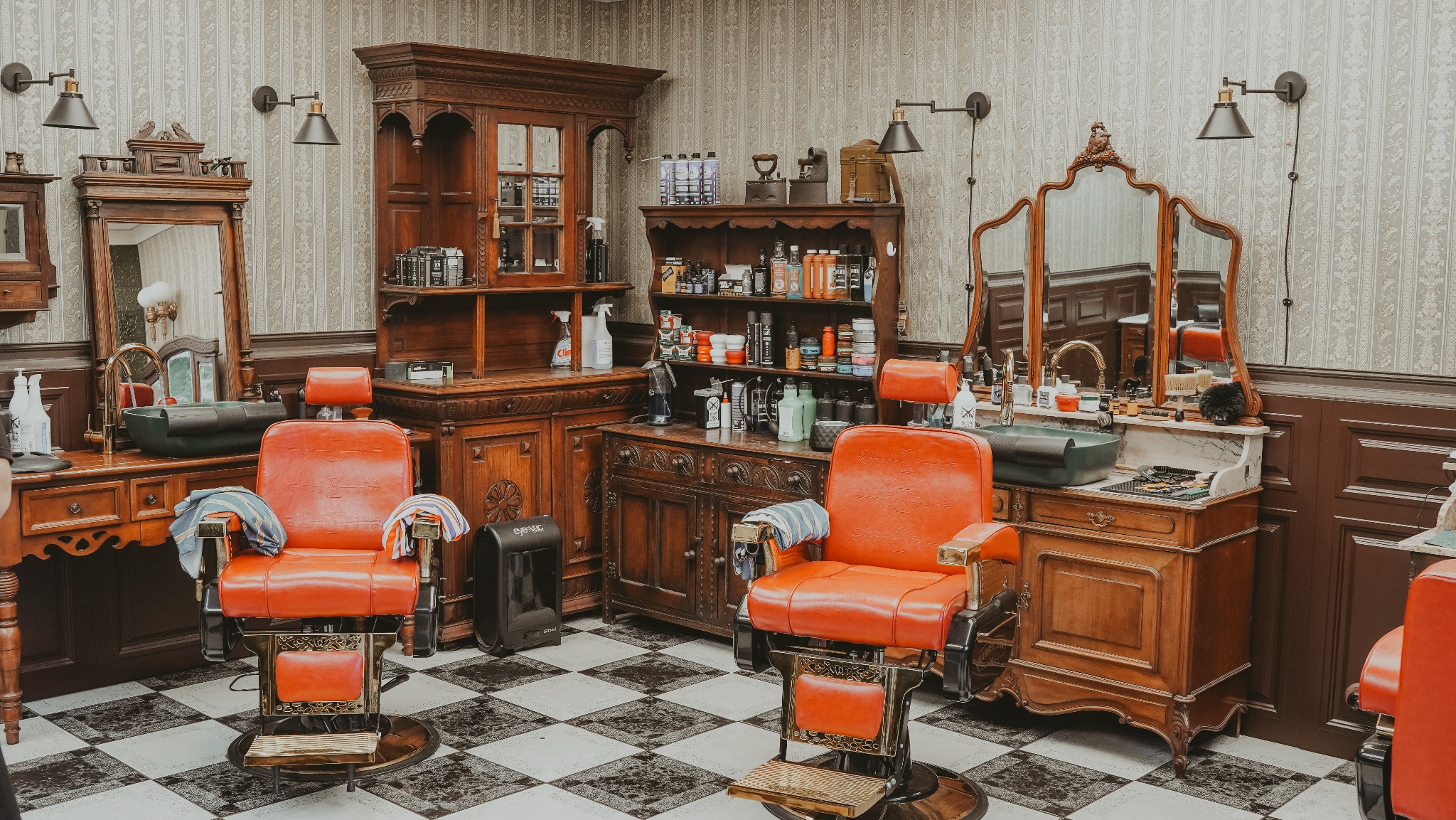 Wnętrze zakładu z dwoma fotelami fryzjerskimi w stylu retro, drewnianymi szafkami i wzorzystą tapetą. Na blacie akcesoria fryzjerskie i umywalka. Czarno-biała podłoga.