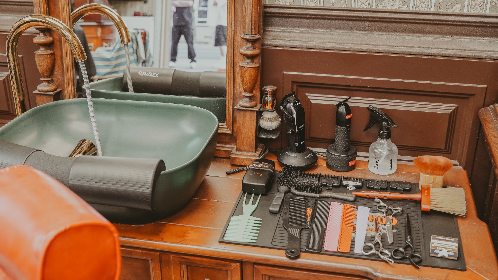 Stanowisko w salonie: umywalka z kranem, maszynki, pędzle, grzebienie i nożyczki na brązowym drewnianym blacie. W tle widoczne lustro i fragment osoby.
