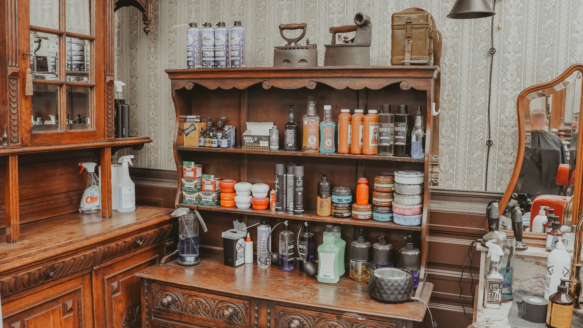 Retro fryzjerski kredens z kosmetykami, narzędziami i zabytkowymi żelazkami. Lustro odbija klienta na fotelu. Tapeta w tle dodaje uroku.