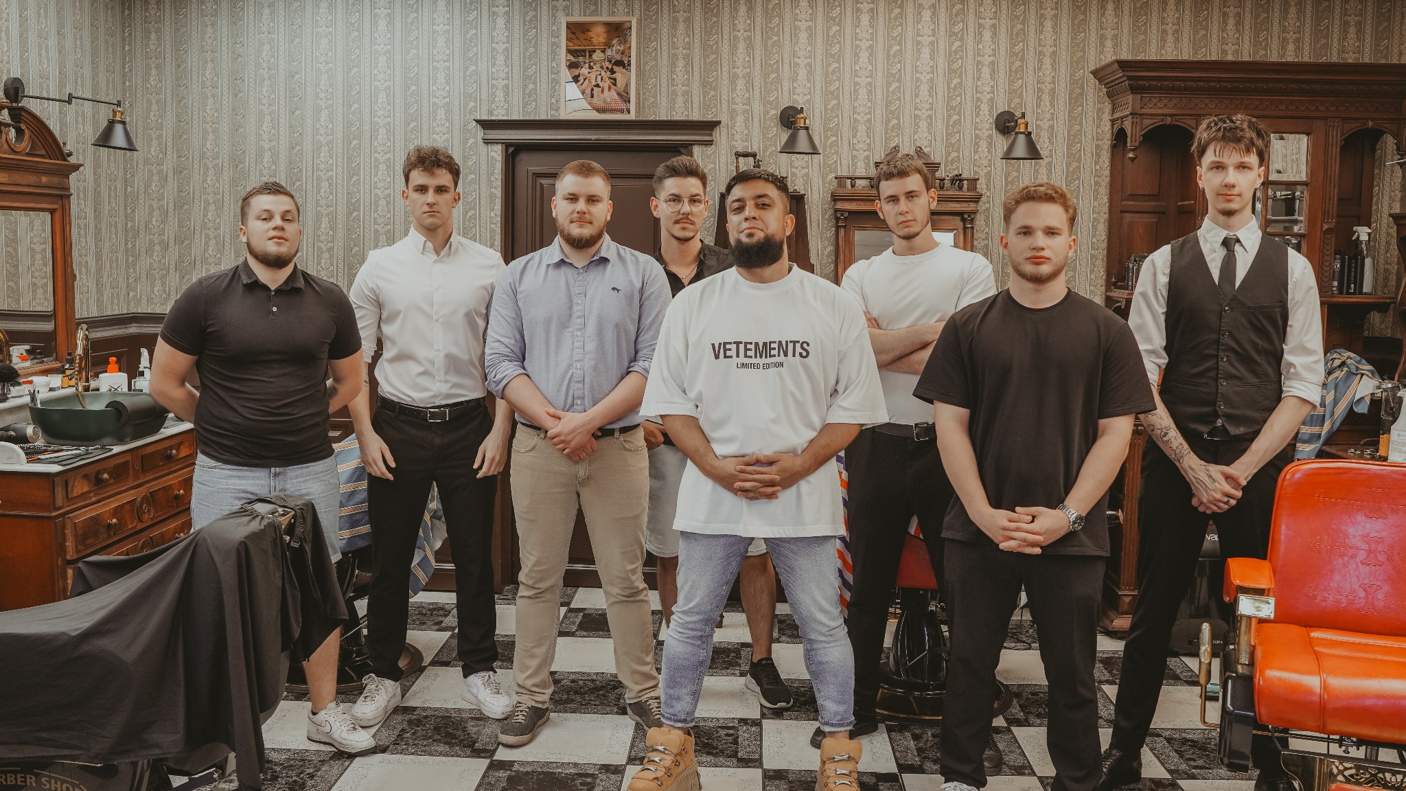 Grupa mężczyzn w barbershopie z tapetą w stylu vintage, prezentujących różne style ubioru. W tle widoczne klasyczne meble i fotel fryzjerski.