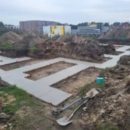 Wylane fundamenty pod budynek z widocznymi deskowaniami i wystającymi elementami. W tle ziemia, palety z bloczkami i inne materiały budowlane.