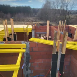 Wznoszenie ścian z czerwonej cegły. Widoczne żółte elementy szalunków i zbrojenia. W tle las i błękitne niebo. Budowa domu w trakcie realizacji.