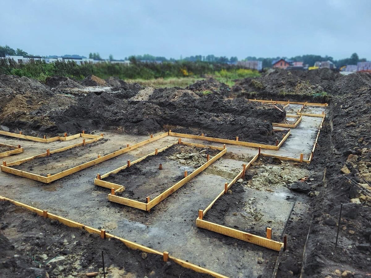 Zalane fundamenty z drewnianym szalunkiem, widoczne wystające pręty zbrojeniowe. Tło: ziemia, trawa, zabudowania. Pochmurne niebo.