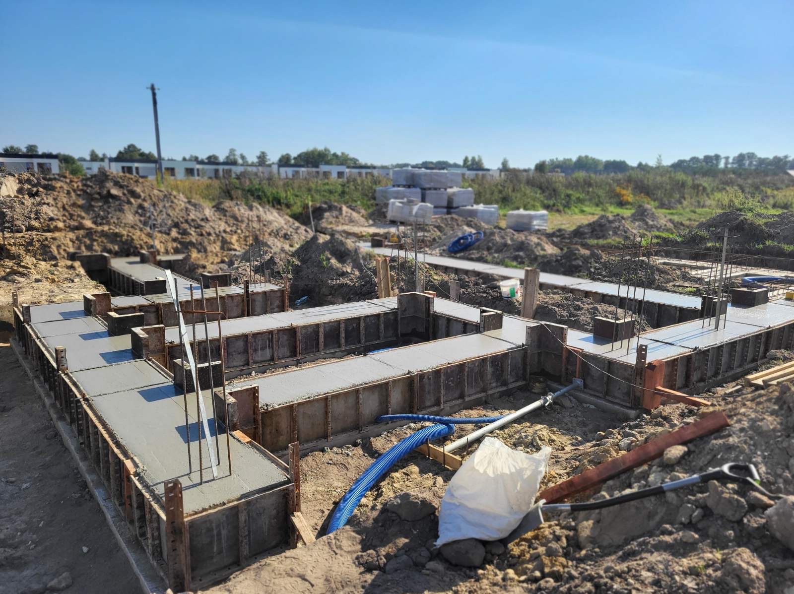Zalane betonem, szalowane fundamenty domu jednorodzinnego. Widoczne zbrojenia i elementy konstrukcyjne. W tle ziemia, budynki w budowie i błękitne niebo.