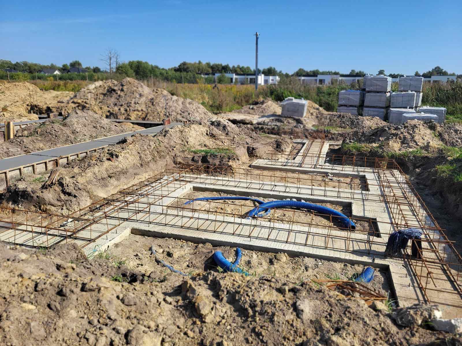 Zbrojenie fundamentów domu jednorodzinnego z widocznymi niebieskimi rurami instalacyjnymi zatopionymi w betonie. W tle ziemia i materiały budowlane.