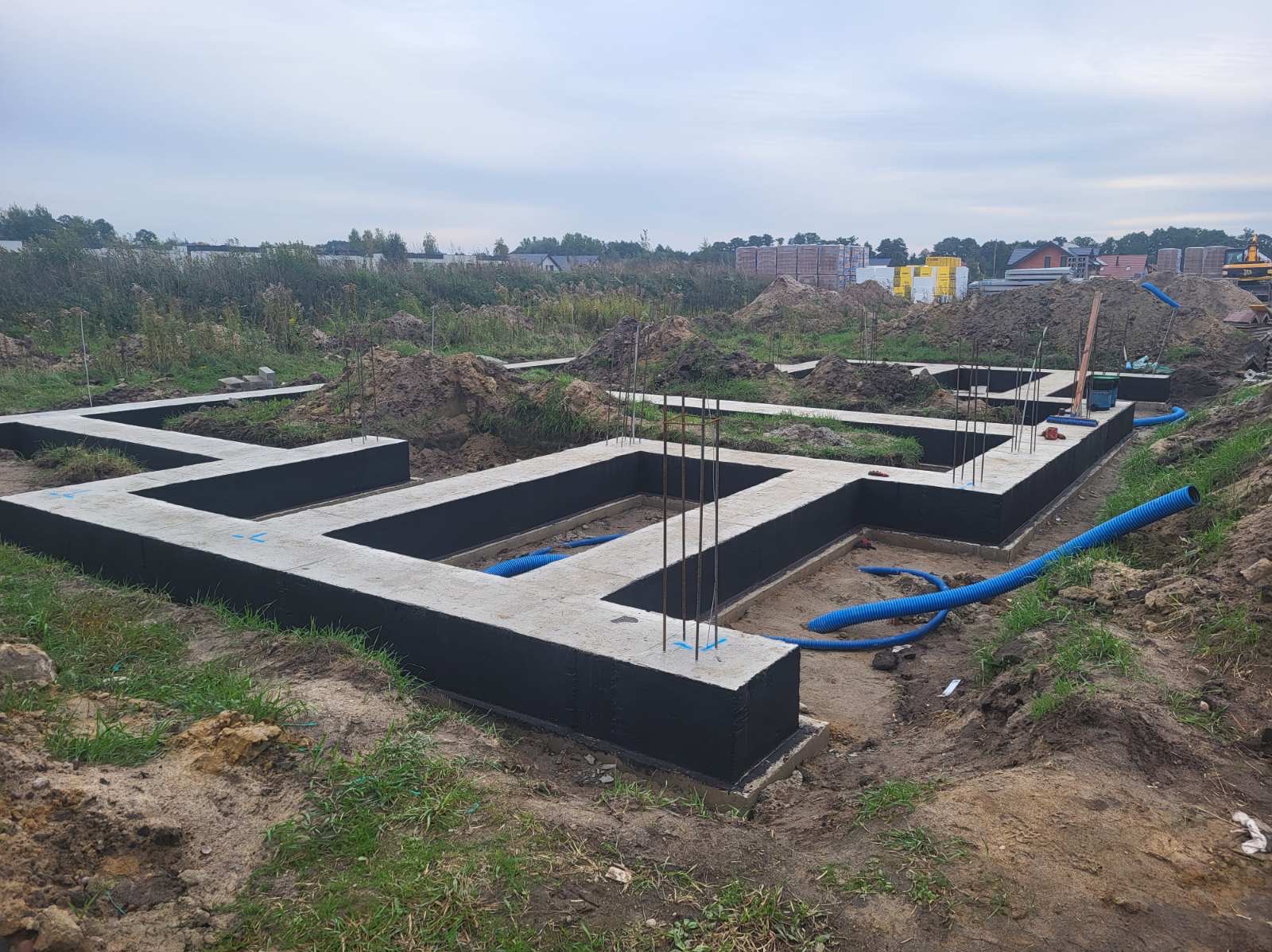 Zalane fundamenty domu jednorodzinnego z izolacją pionową, widoczne zbrojenia i niebieskie rury drenażowe, ziemia, trawa, w tle zabudowa i hałda materiałów budowlanych.
