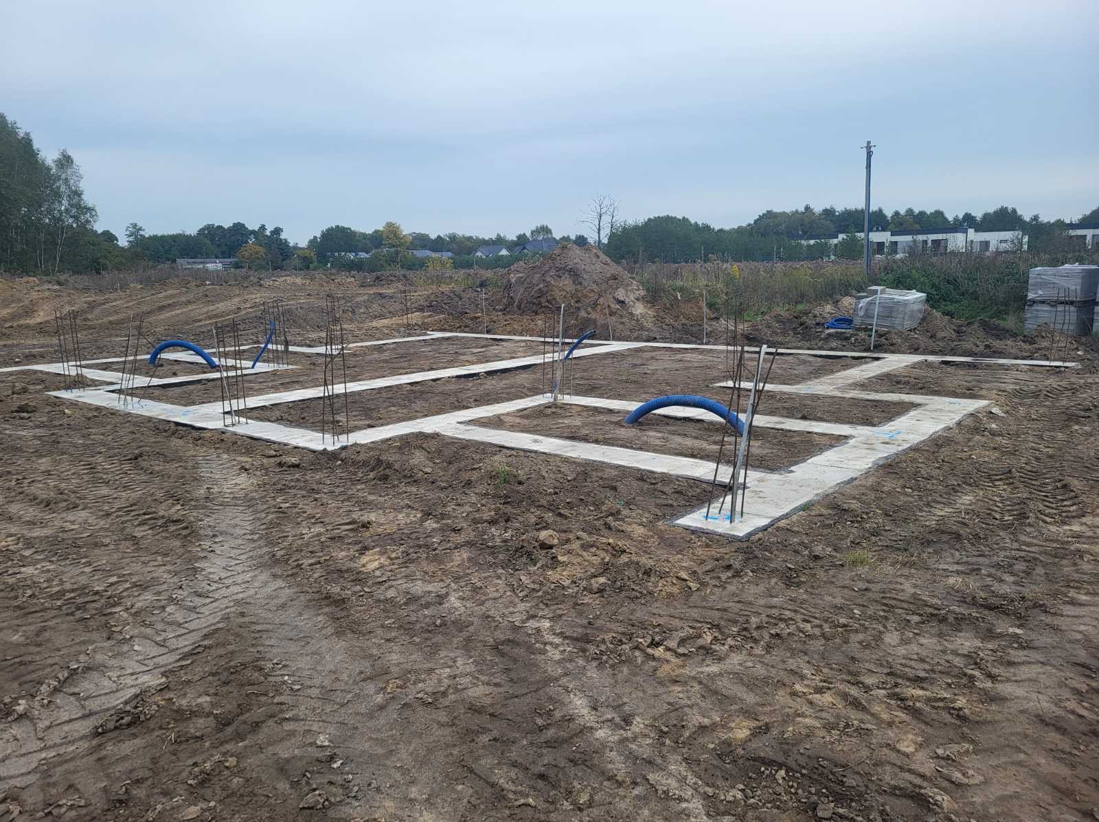 Zalane fundamenty domu jednorodzinnego z wystającymi prętami zbrojeniowymi i niebieskimi rurami na gruncie. Widok z góry na plac budowy w pochmurny dzień.