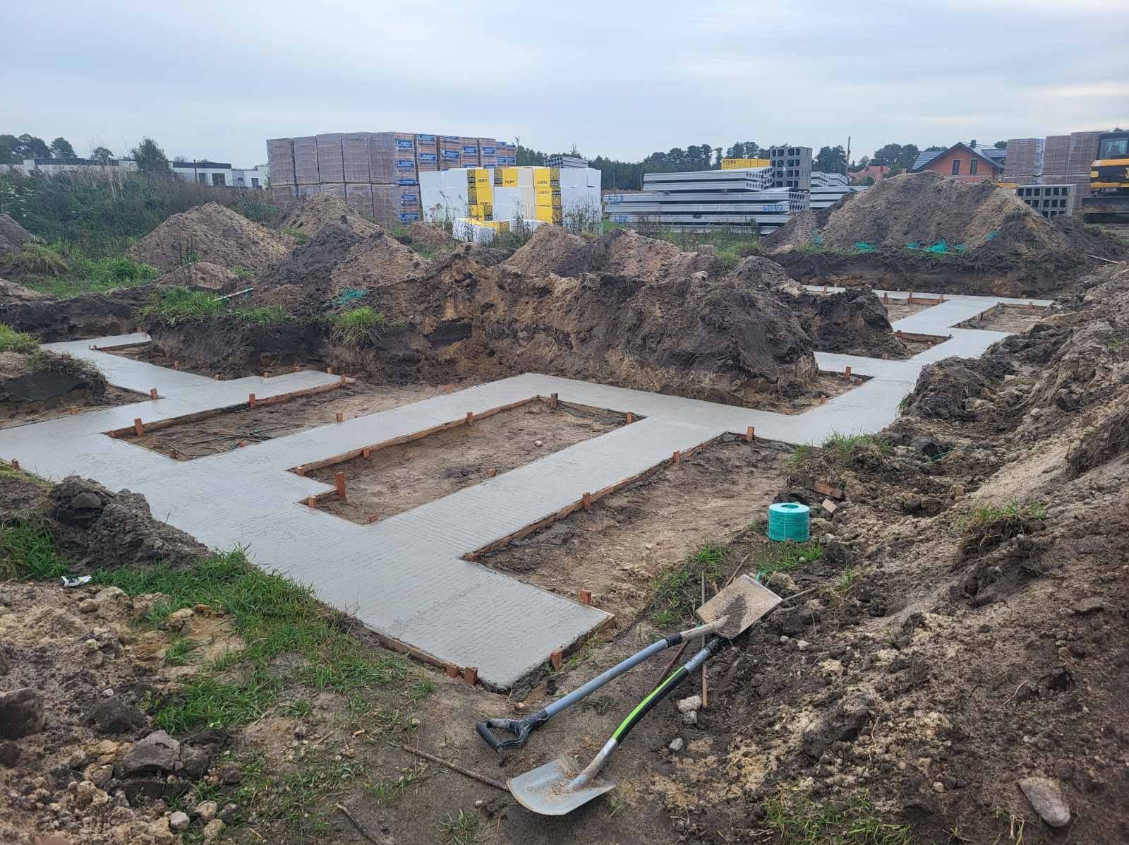 Wylane fundamenty pod budynek z widocznymi deskowaniami i wystającymi elementami. W tle ziemia, palety z bloczkami i inne materiały budowlane.