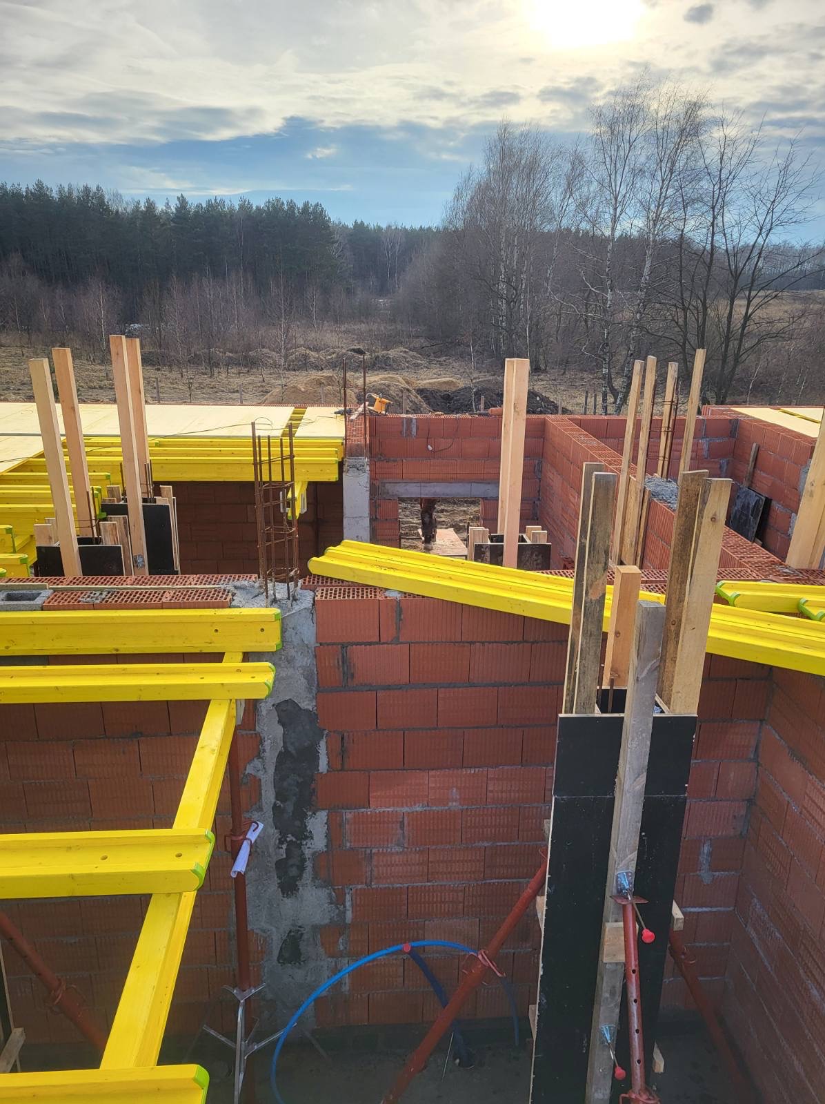 Wznoszenie ścian z czerwonej cegły. Widoczne żółte elementy szalunków i zbrojenia. W tle las i błękitne niebo. Budowa domu w trakcie realizacji.