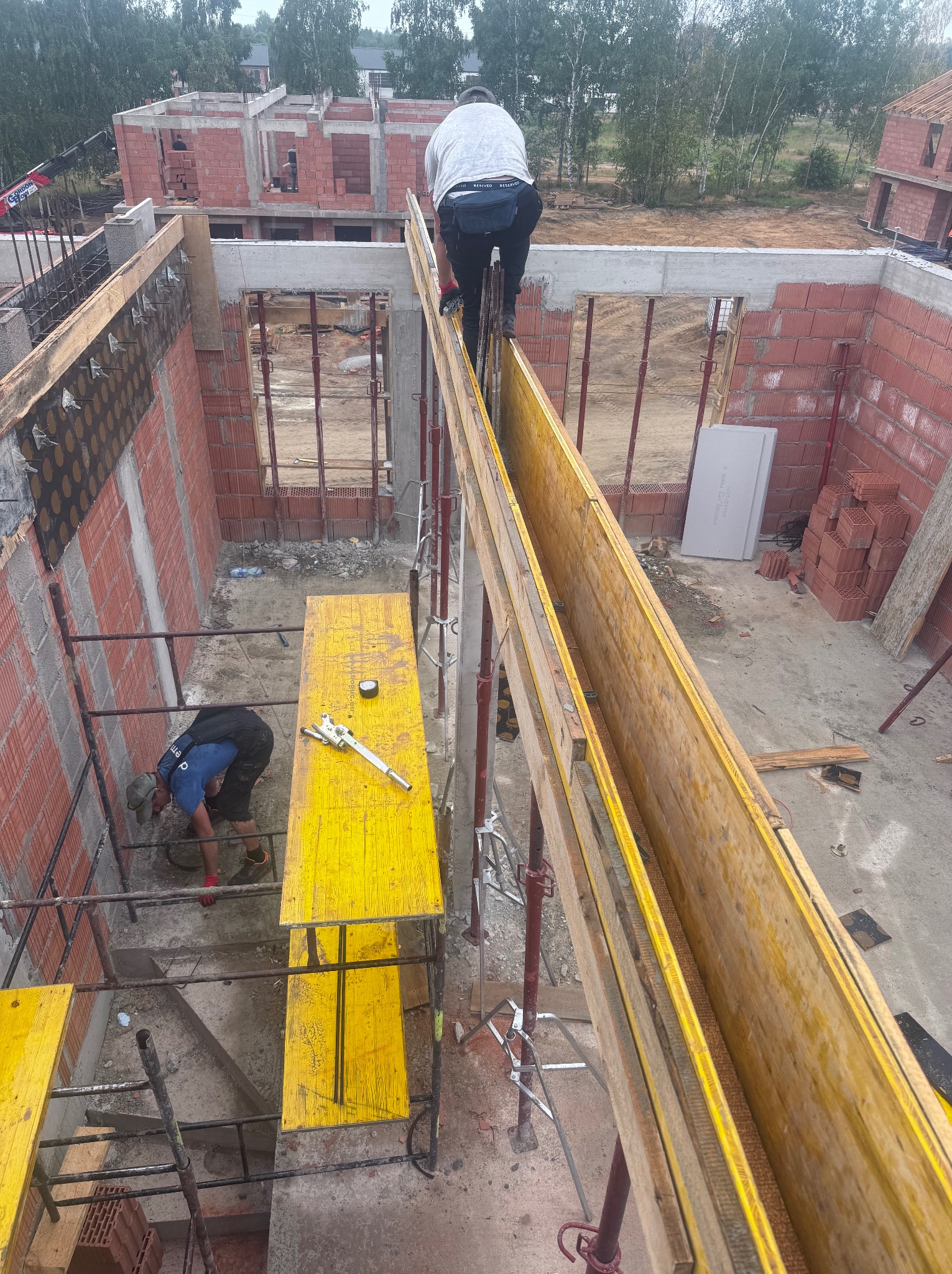 Budowa domu w trakcie: pracownicy na rusztowaniach, szalunki, murowane ściany z czerwonej cegły, widok z góry. Etap prac budowlanych.