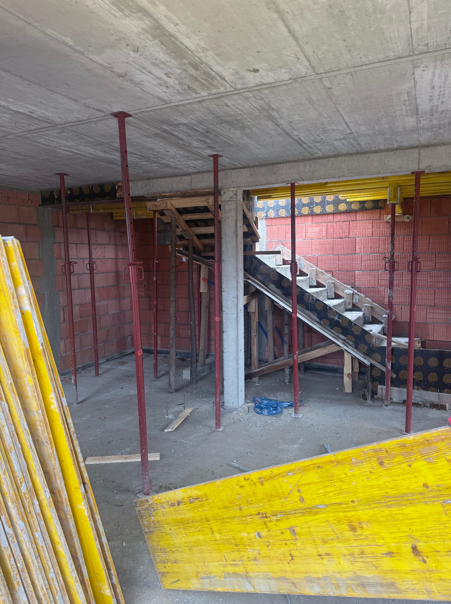 Wnętrze w trakcie budowy: czerwona cegła, szalunki schodów z czarnym wzorem, podpory stropowe i żółte deski szalunkowe, surowy betonowy strop.