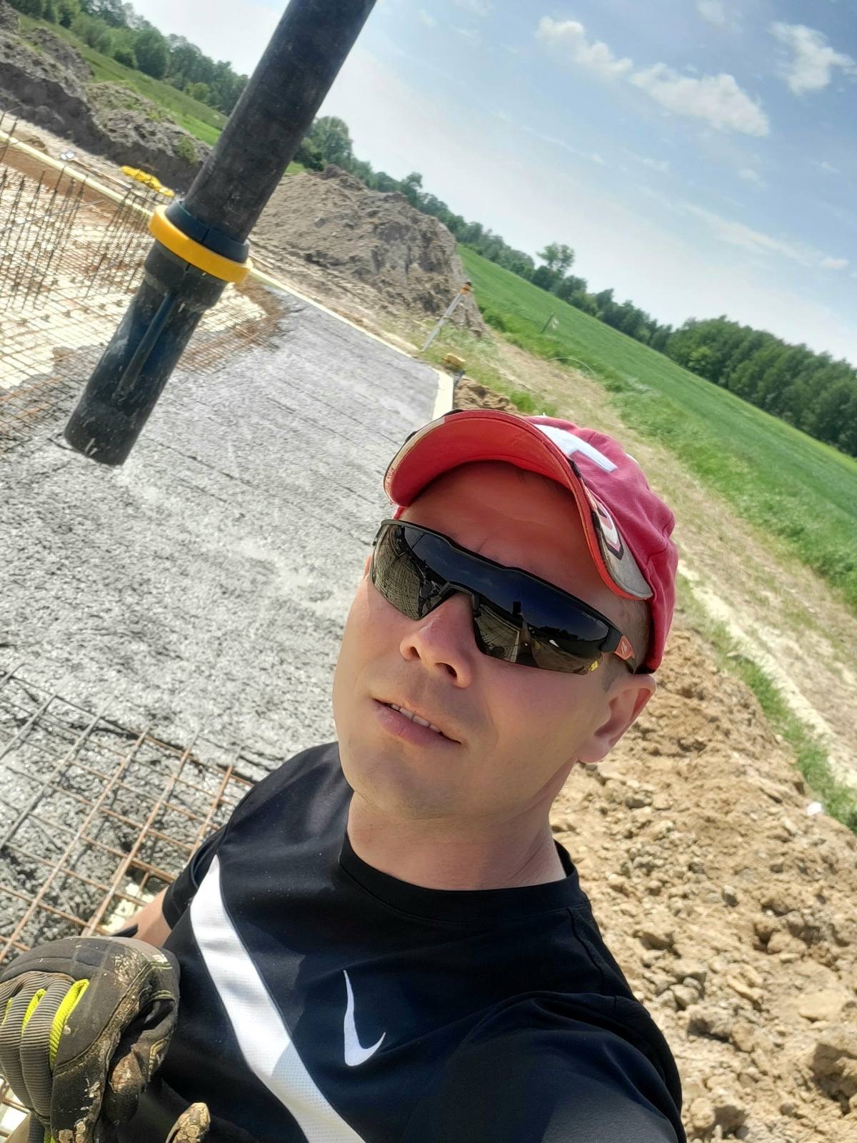 Wylany beton na zbrojeniu fundamentów domu jednorodzinnego. W tle krajobraz polny i las. Widoczny pracownik w okularach i czapce.