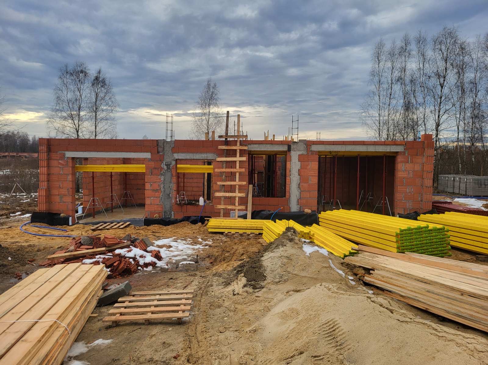Wznoszenie ścian domu z czerwonej cegły, widoczne żółte podpory stropowe, drewniane deski i piasek. Prace budowlane w toku, pochmurne niebo w tle.
