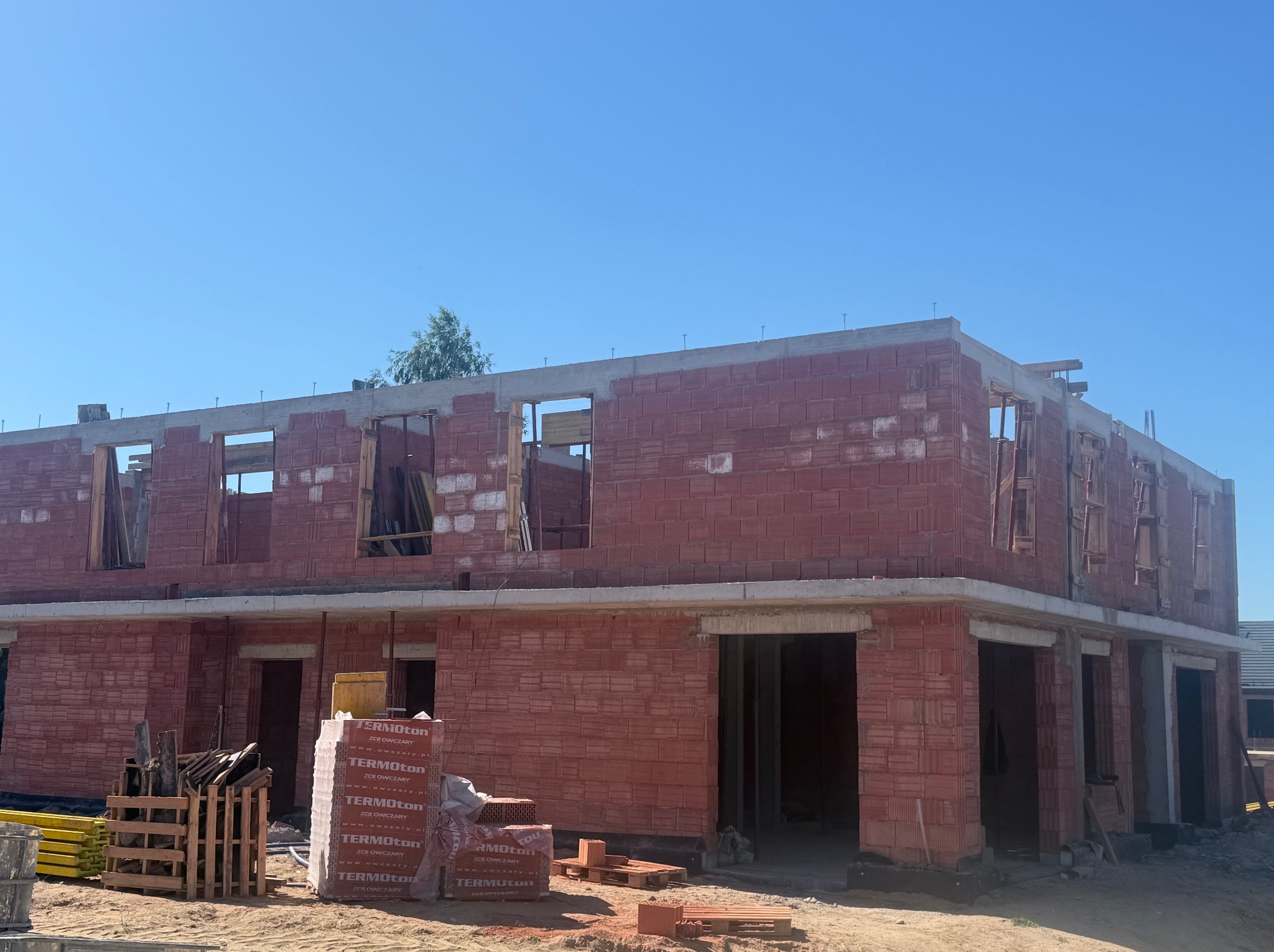 Wznoszenie dwukondygnacyjnego domu z czerwonej cegły Termoton. Widoczne pakiety cegieł, pustaki i przygotowane otwory okienne. Słoneczny dzień, błękitne niebo.