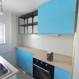 RM-PRODUKCJA RAFAŁ MONDEL - Błękitna zabudowa kuchenna z piekarnikiem i drewnianym blatem. Na blacie kartonowe opakowania. Nad blatem półki i białe płytki imitujące marmur.