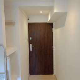 RM-PRODUKCJA RAFAŁ MONDEL - Wnętrze korytarza po remoncie: brązowe drzwi wejściowe z srebrną klamką, świeżo pomalowane białe ściany, jasna podłoga i sufit z oświetleniem punktowym.