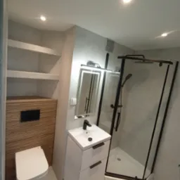 Nowoczesna łazienka z kabiną prysznicową typu walk-in, umywalką z szafką i WC w zabudowie. Minimalistyczny design, szare płytki, czarne akcenty.