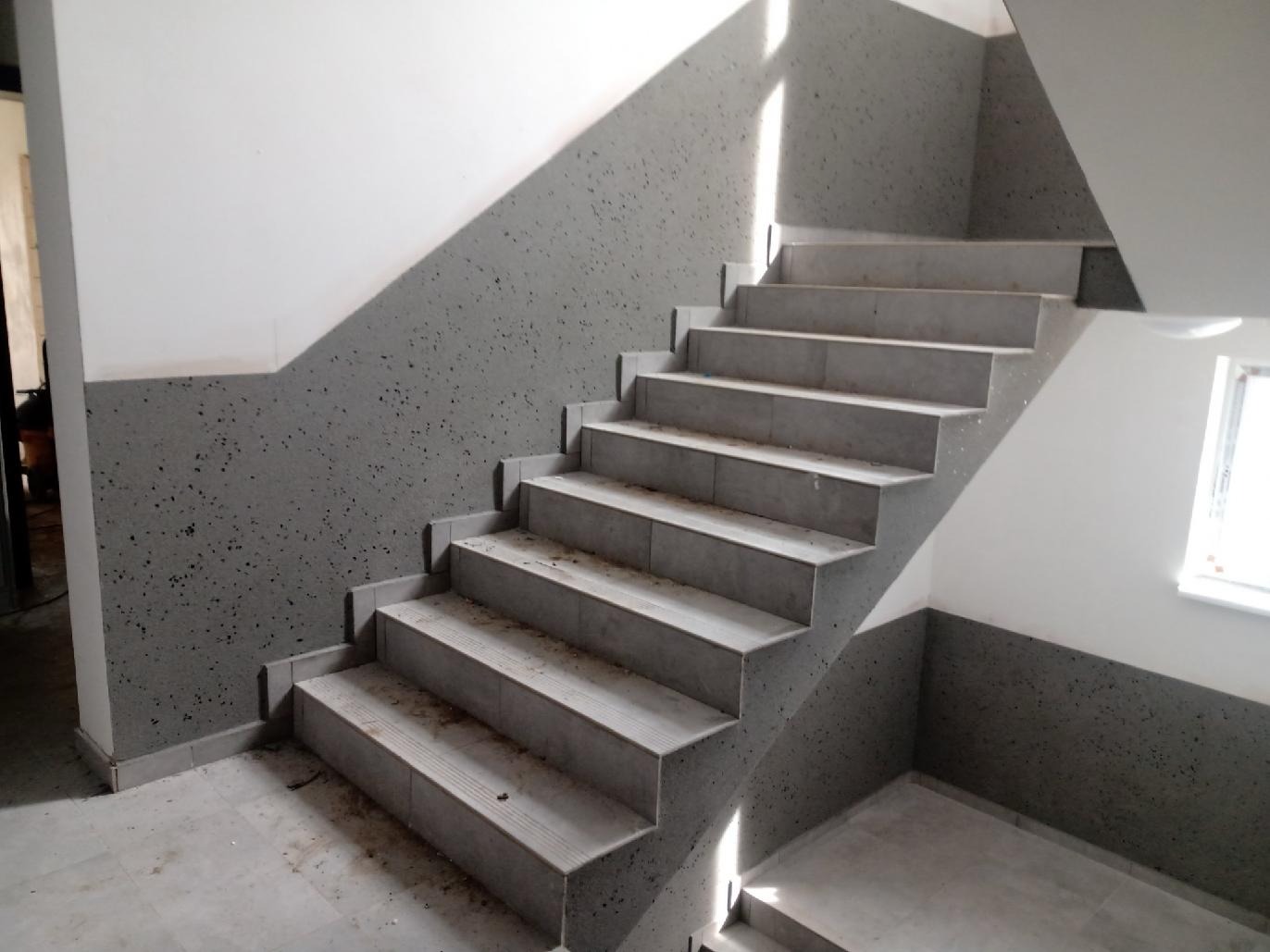 Klatka schodowa po remoncie: szare płytki na schodach, ściany pomalowane na biało i szaro, z charakterystyczną, nakrapianą fakturą szarej farby. Widoczne okno.