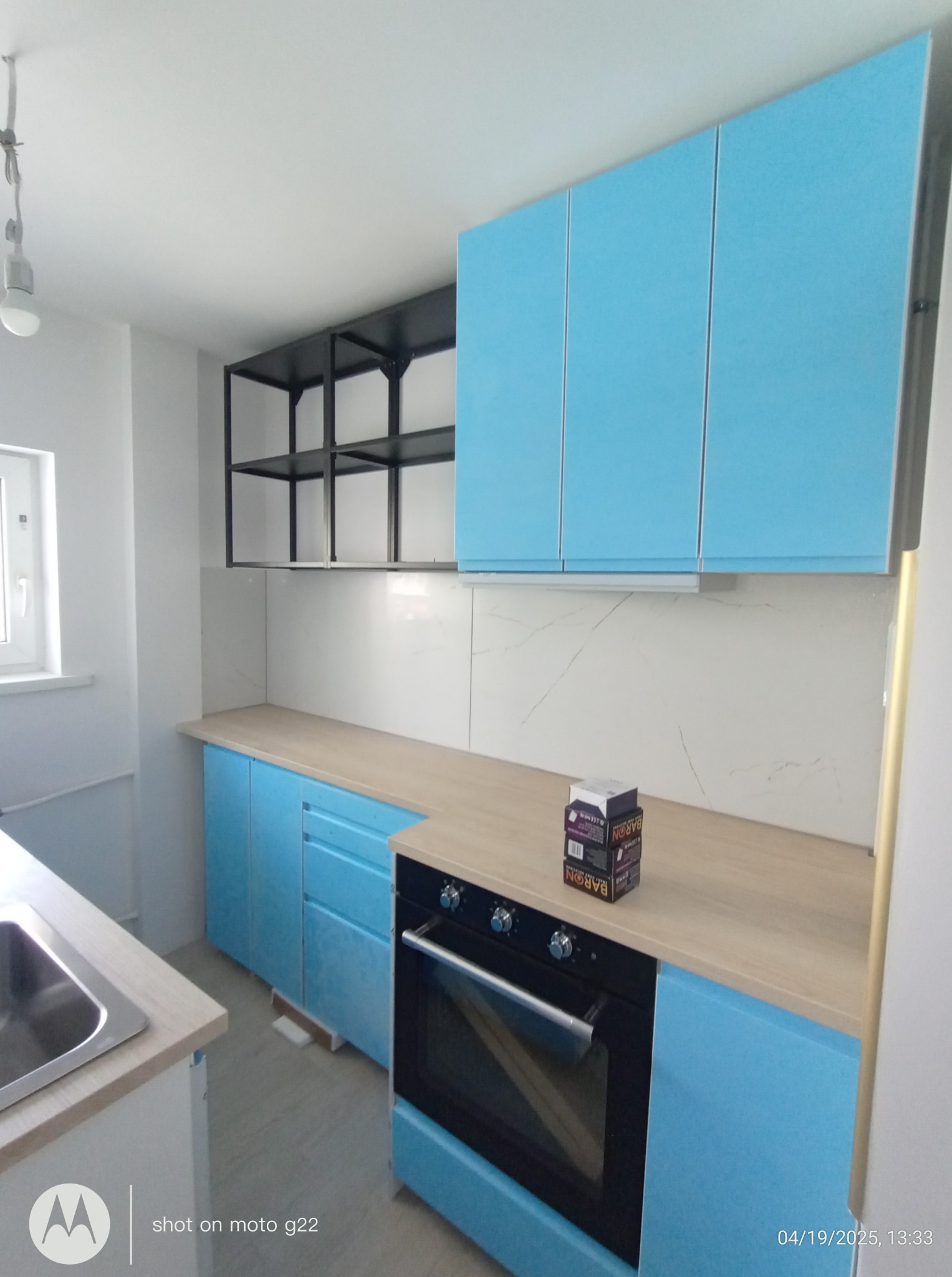 Błękitna zabudowa kuchenna z piekarnikiem i drewnianym blatem. Na blacie kartonowe opakowania. Nad blatem półki i białe płytki imitujące marmur.