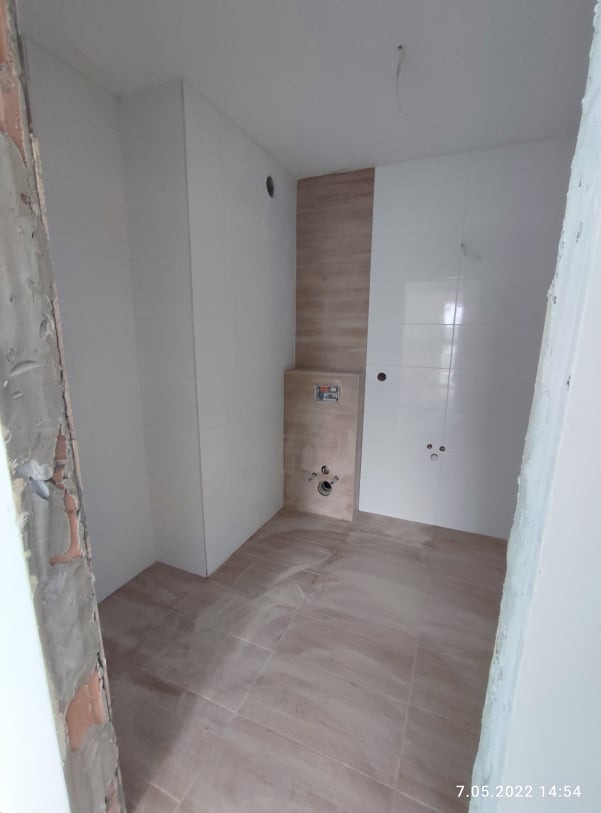 Wnętrze łazienki w trakcie wykończenia. Ułożone beżowe płytki na podłodze i częściowo na ścianach. Widoczne przygotowane instalacje. Surowy fragment ściany przy wejściu.