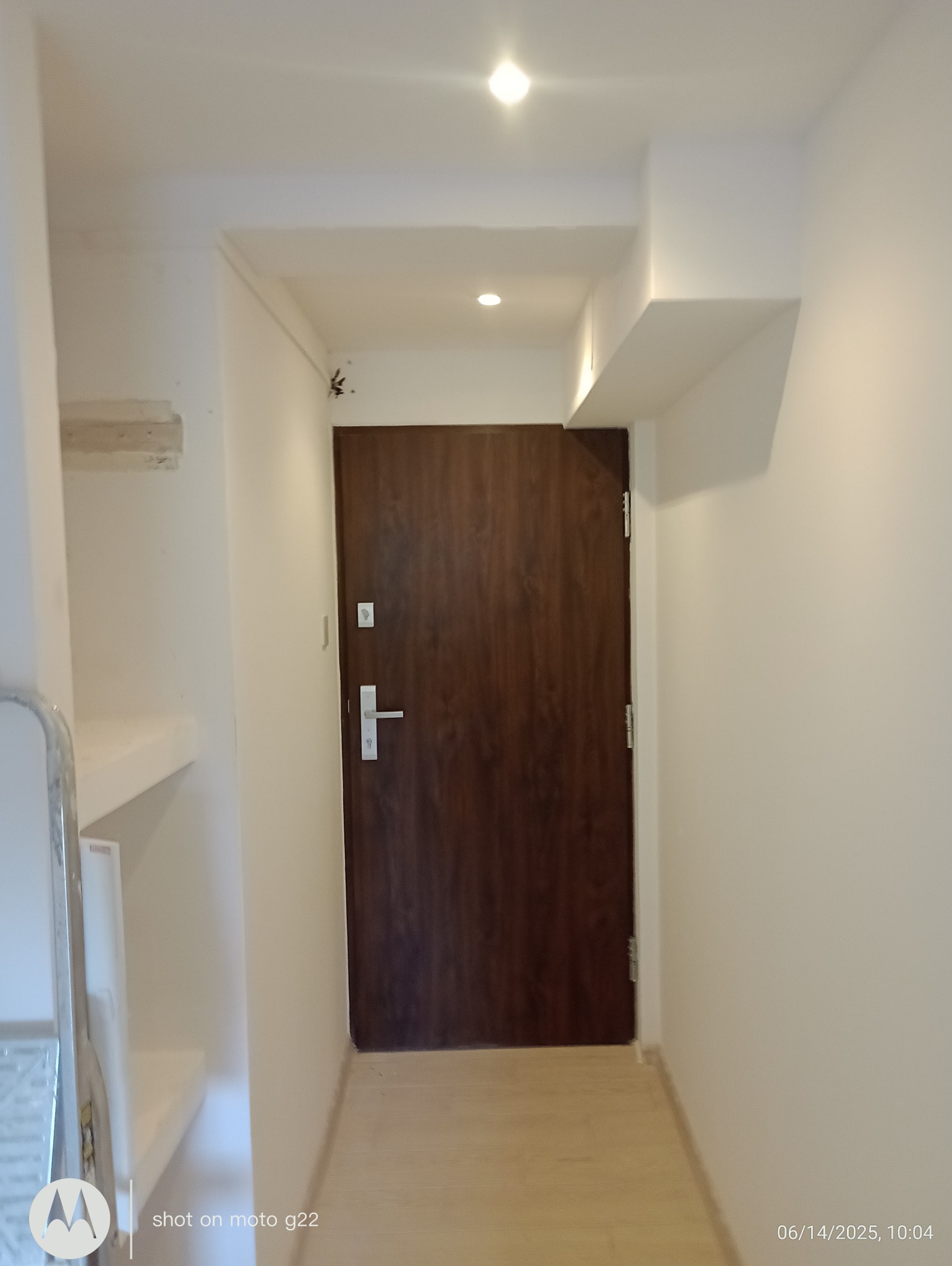 Wnętrze korytarza po remoncie: brązowe drzwi wejściowe z srebrną klamką, świeżo pomalowane białe ściany, jasna podłoga i sufit z oświetleniem punktowym.