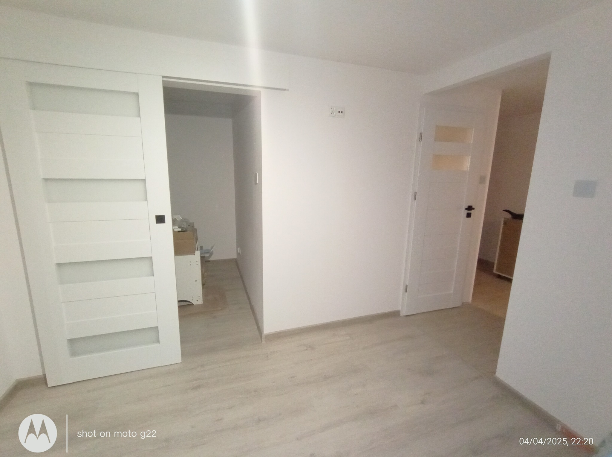 Jasne wnętrze po remoncie: białe ściany, panele podłogowe. Widoczne białe drzwi przesuwne z szybami i standardowe drzwi z klamką. Minimalistyczny, nowoczesny styl.