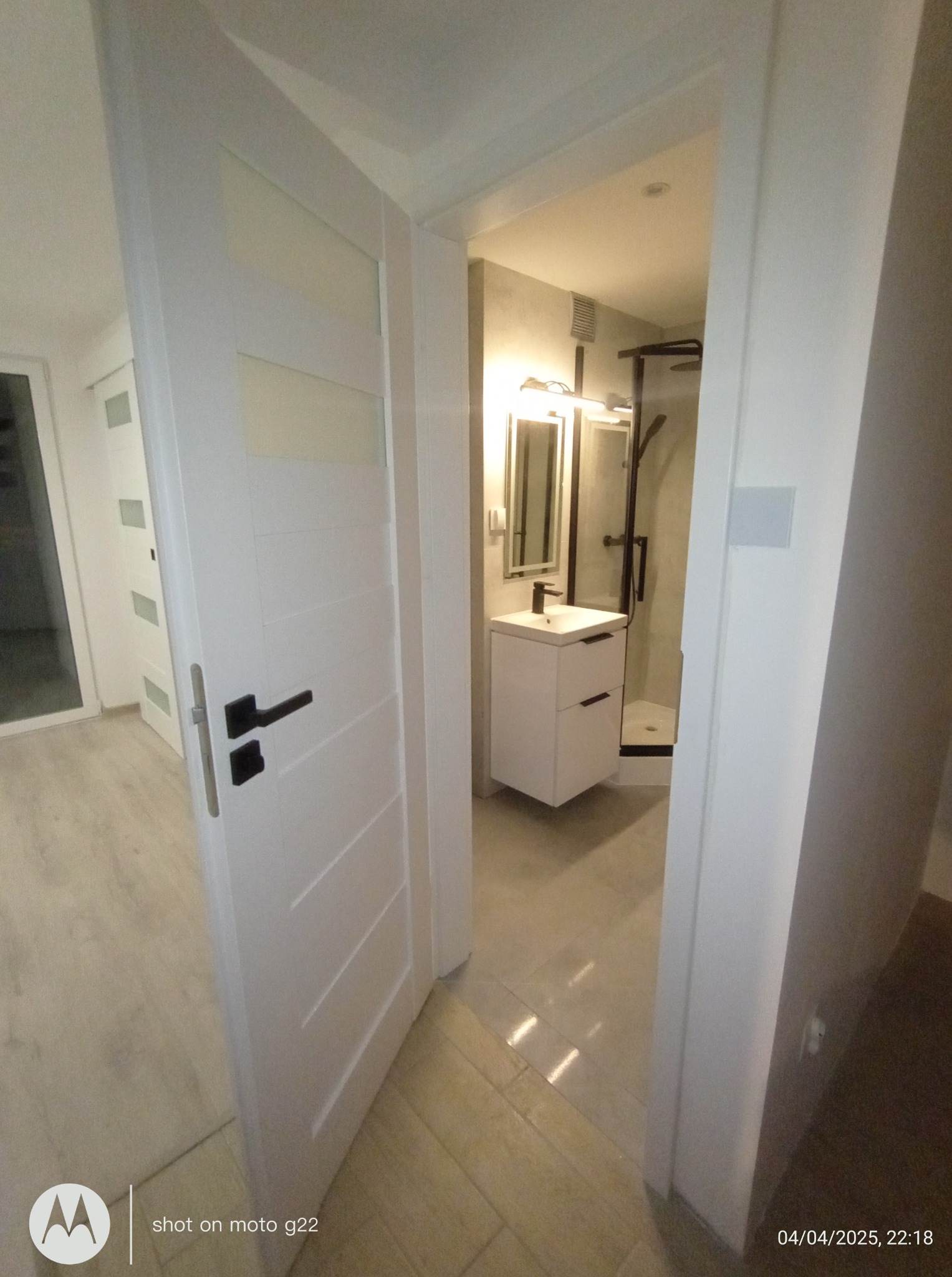 Otwarta, biała, nowoczesna łazienka z kabiną prysznicową i szafką z umywalką, widoczna przez uchylone drzwi z czarną klamką. Jasne płytki podłogowe.