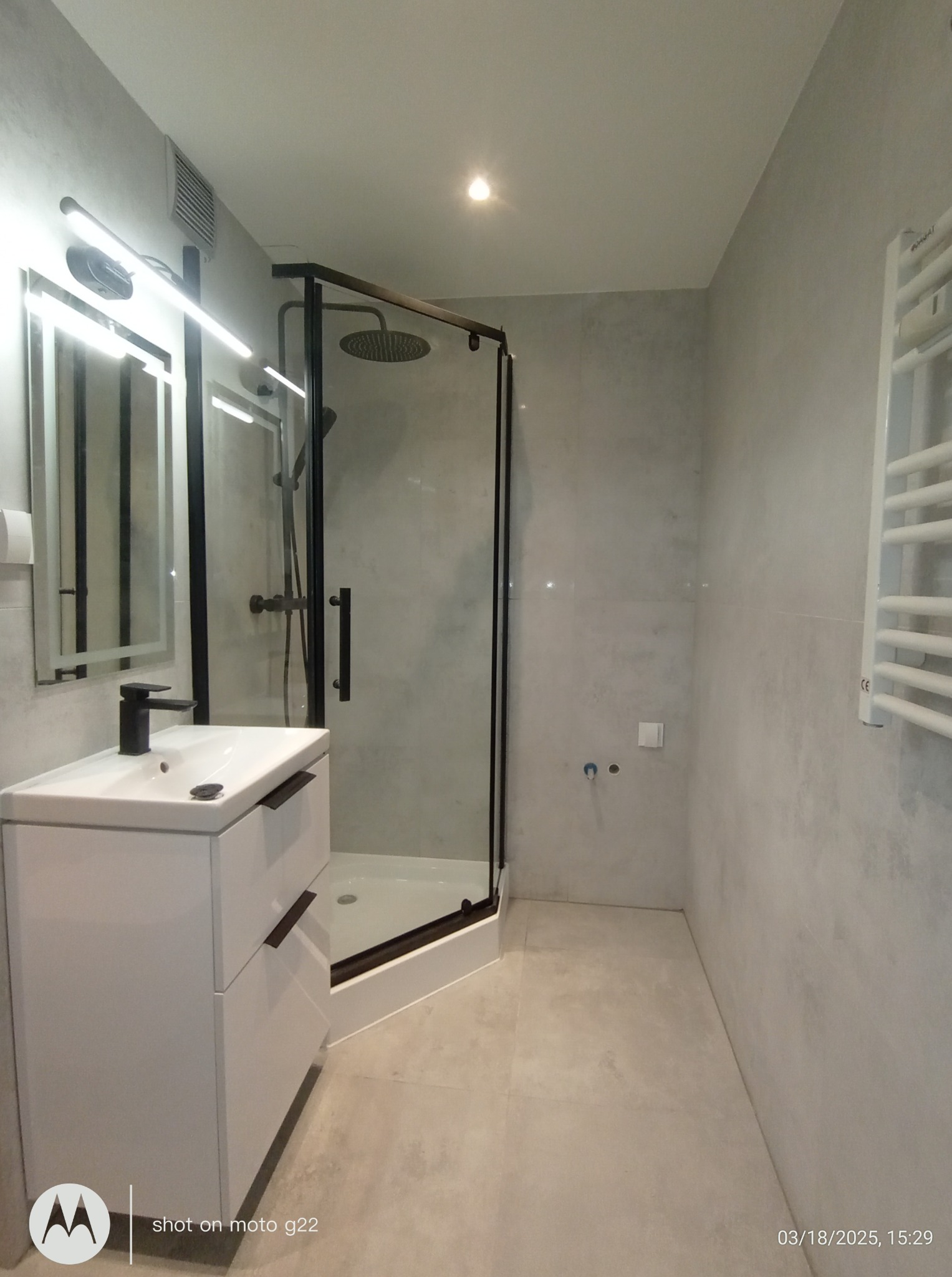 Nowoczesna łazienka z kabiną prysznicową typu walk-in z czarnymi profilami, białą szafką pod umywalką i grzejnikiem drabinkowym. Minimalistyczny design i szara kolorystyka.