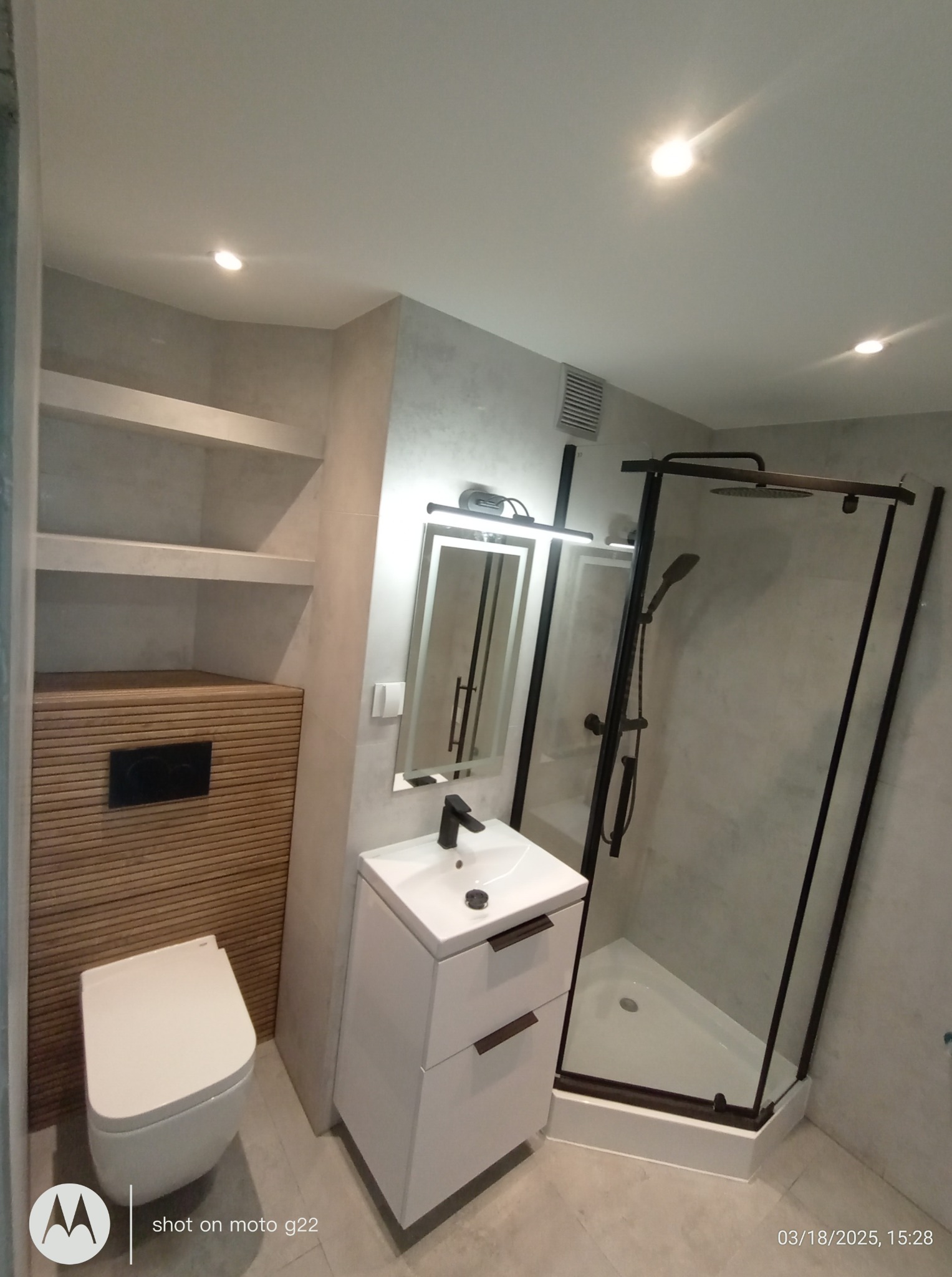 Nowoczesna łazienka z kabiną prysznicową typu walk-in, umywalką z szafką i WC w zabudowie. Minimalistyczny design, szare płytki, czarne akcenty.