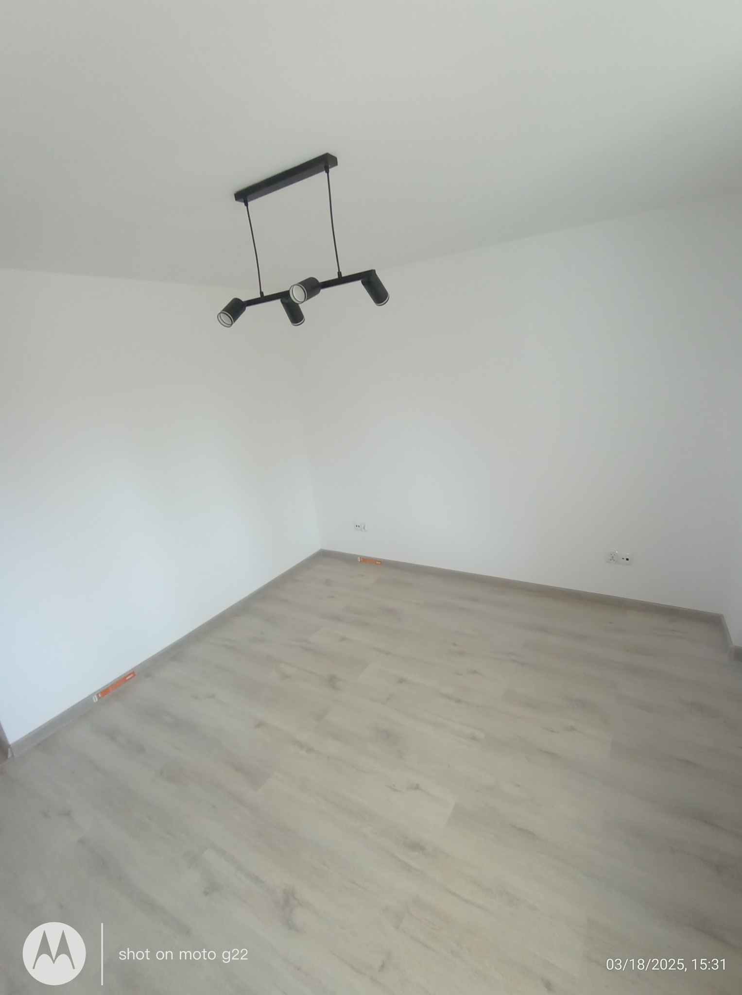 Jasne panele podłogowe w pustym pokoju z białymi ścianami i nowoczesną, czarną lampą sufitową. Widoczny fragment listwy przypodłogowej.