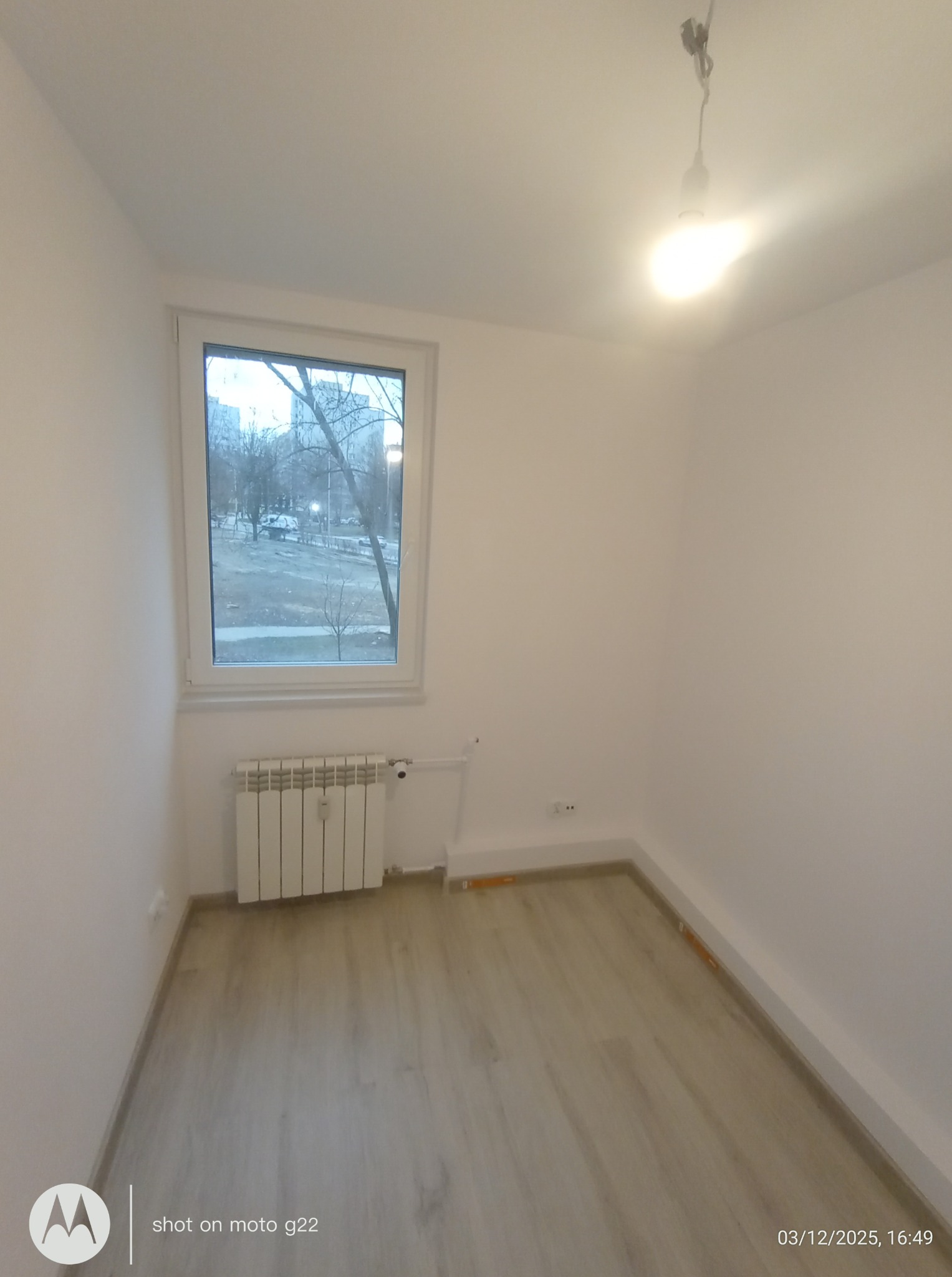 Pusty pokój po remoncie: świeżo pomalowane ściany na biało, jasna podłoga, grzejnik pod oknem z widokiem na drzewa. Minimalizm i gotowość do aranżacji.