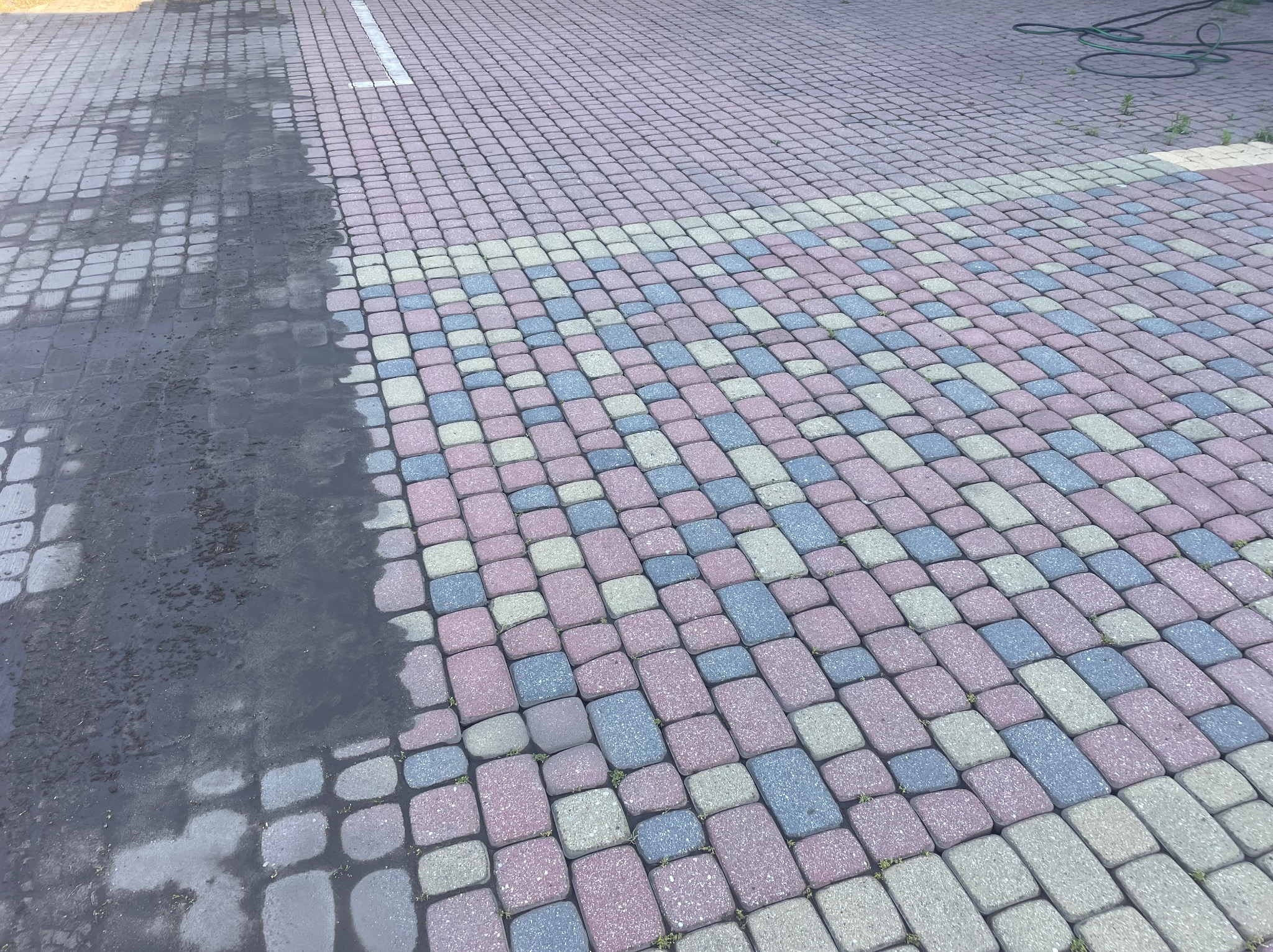 Częściowo umyta, kolorowa kostka brukowa. Widoczny kontrast między czystą, jasną powierzchnią a brudną, ciemną. W tle wąż ogrodowy.