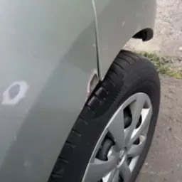 Fragment srebrnego auta z widocznymi uszkodzeniami karoserii i śladami szpachlowania przy kole. Widoczny fragment opony i felgi.